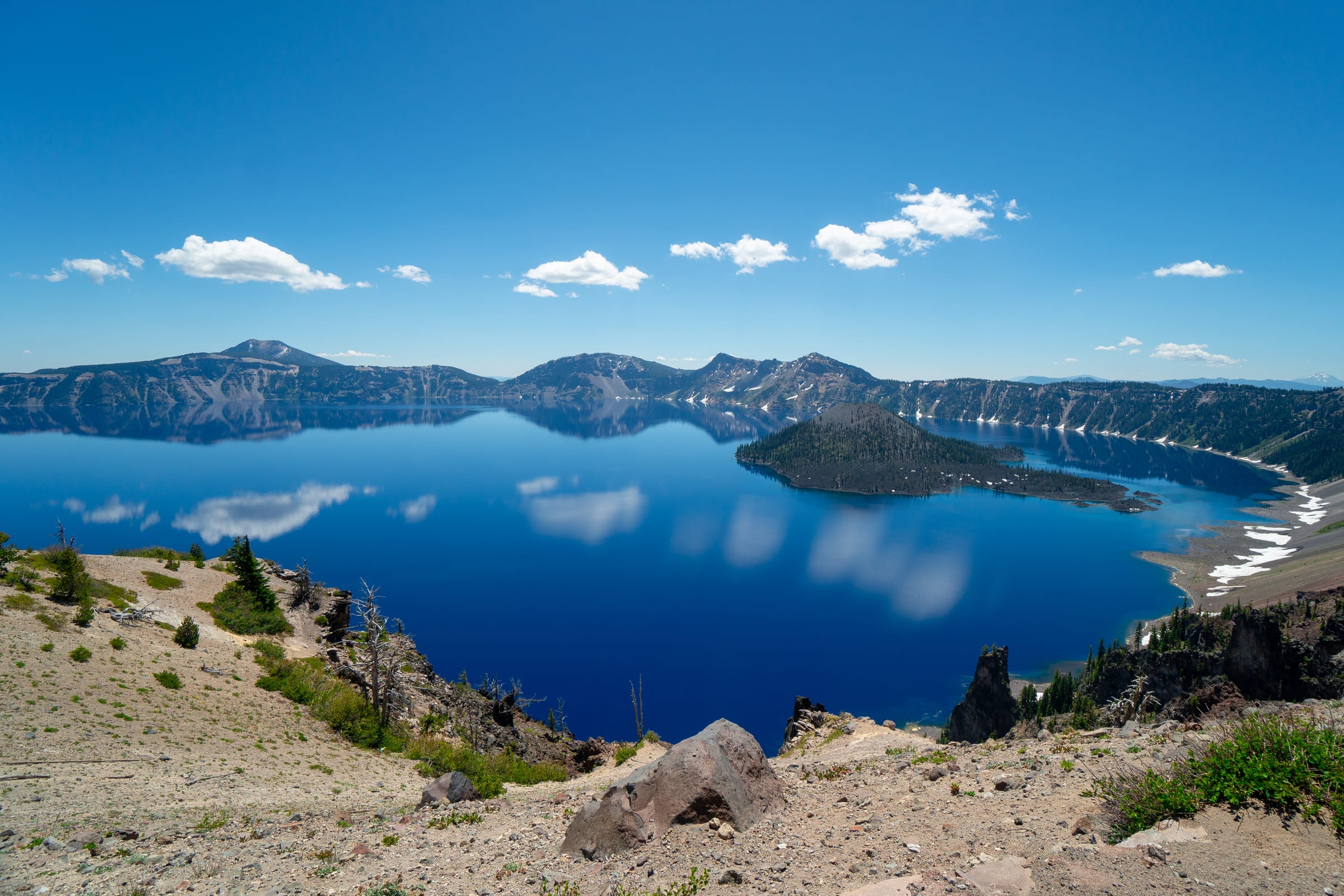 Wizard Island in the deep blue water of Crater Lake in July, clouds reflected on the surface