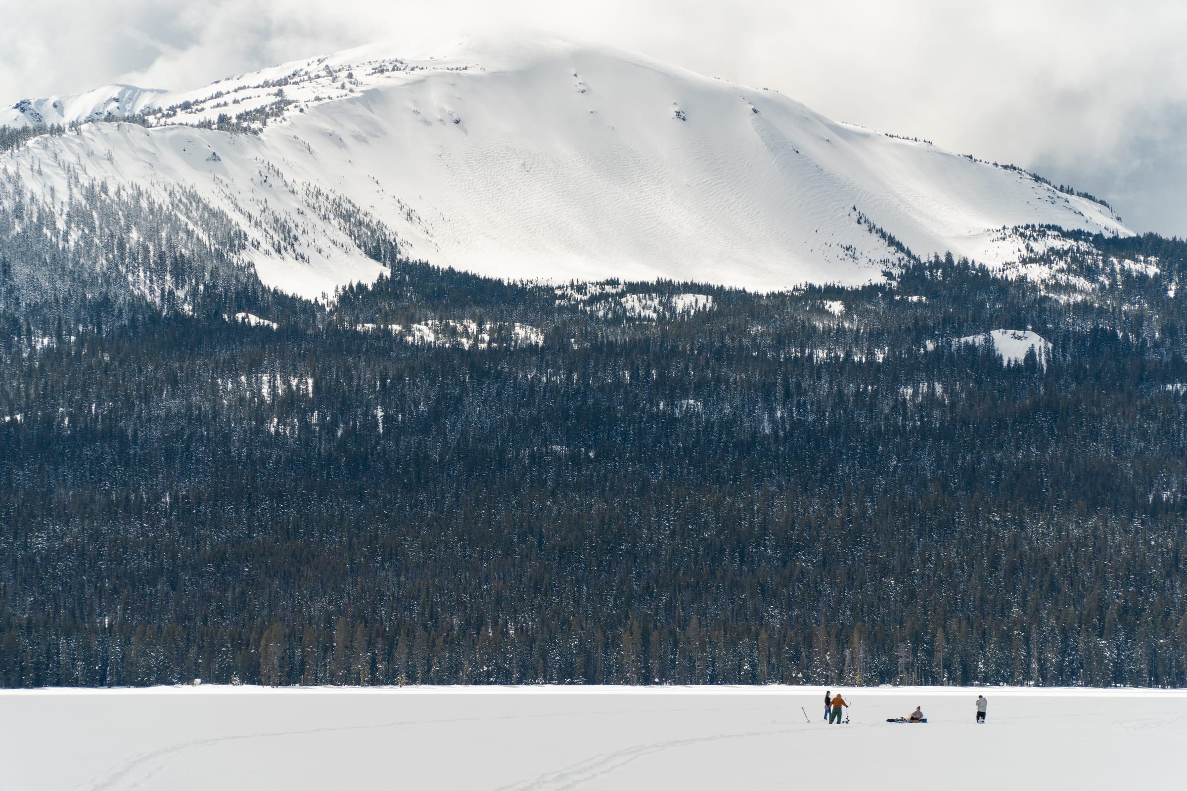 People ice fishing on frozen Diamond Lake in March under a snow-covered mountain and dark forest