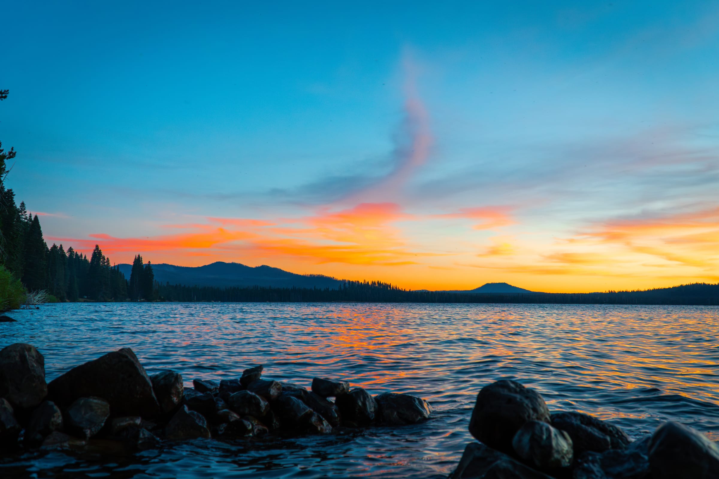 An orange and blue August sunset over Diamond Lake, the peak of Mount Thielsen dark against the sky and rocks at the water's edge