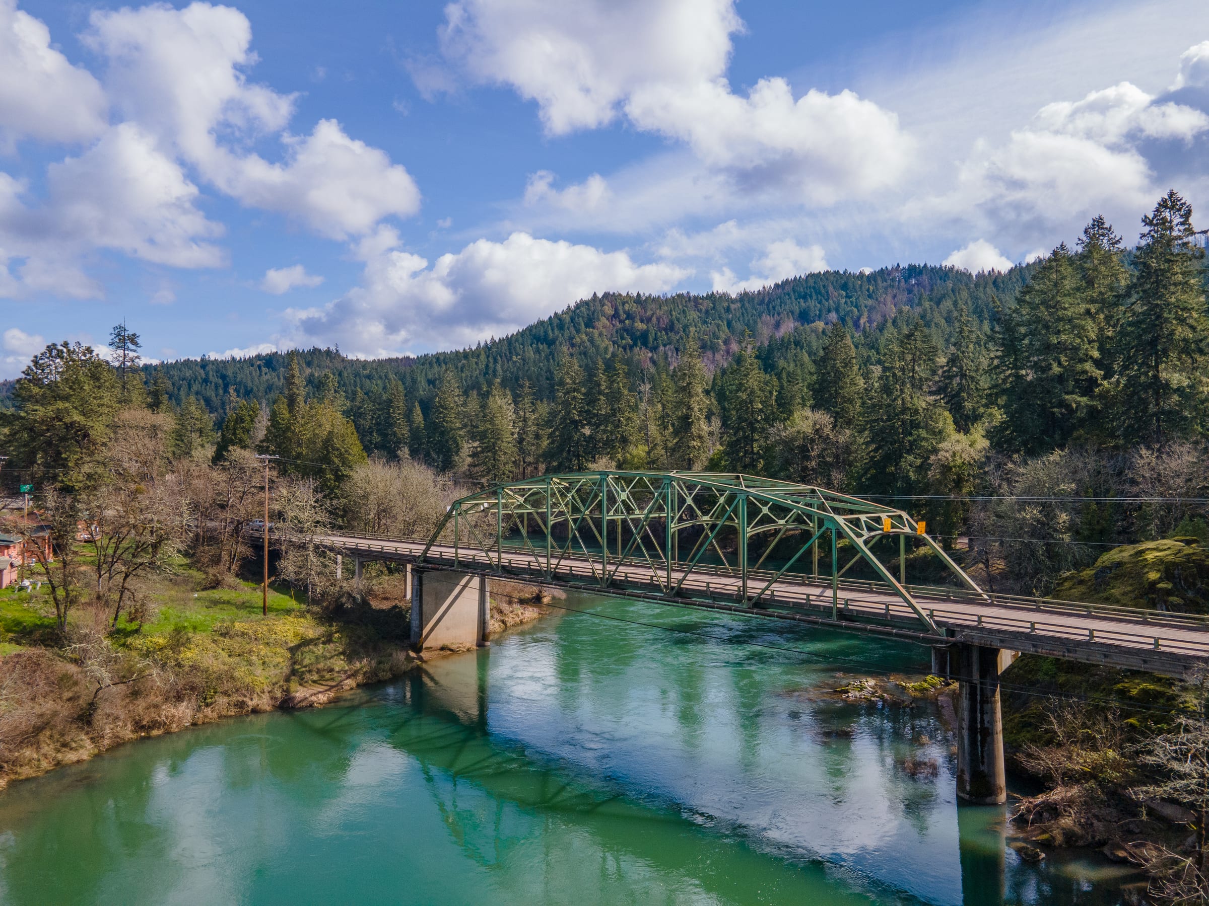 A green steel bridge over the clear turquoise North Umpqua River at Glide in March, forested hills and white clouds behind