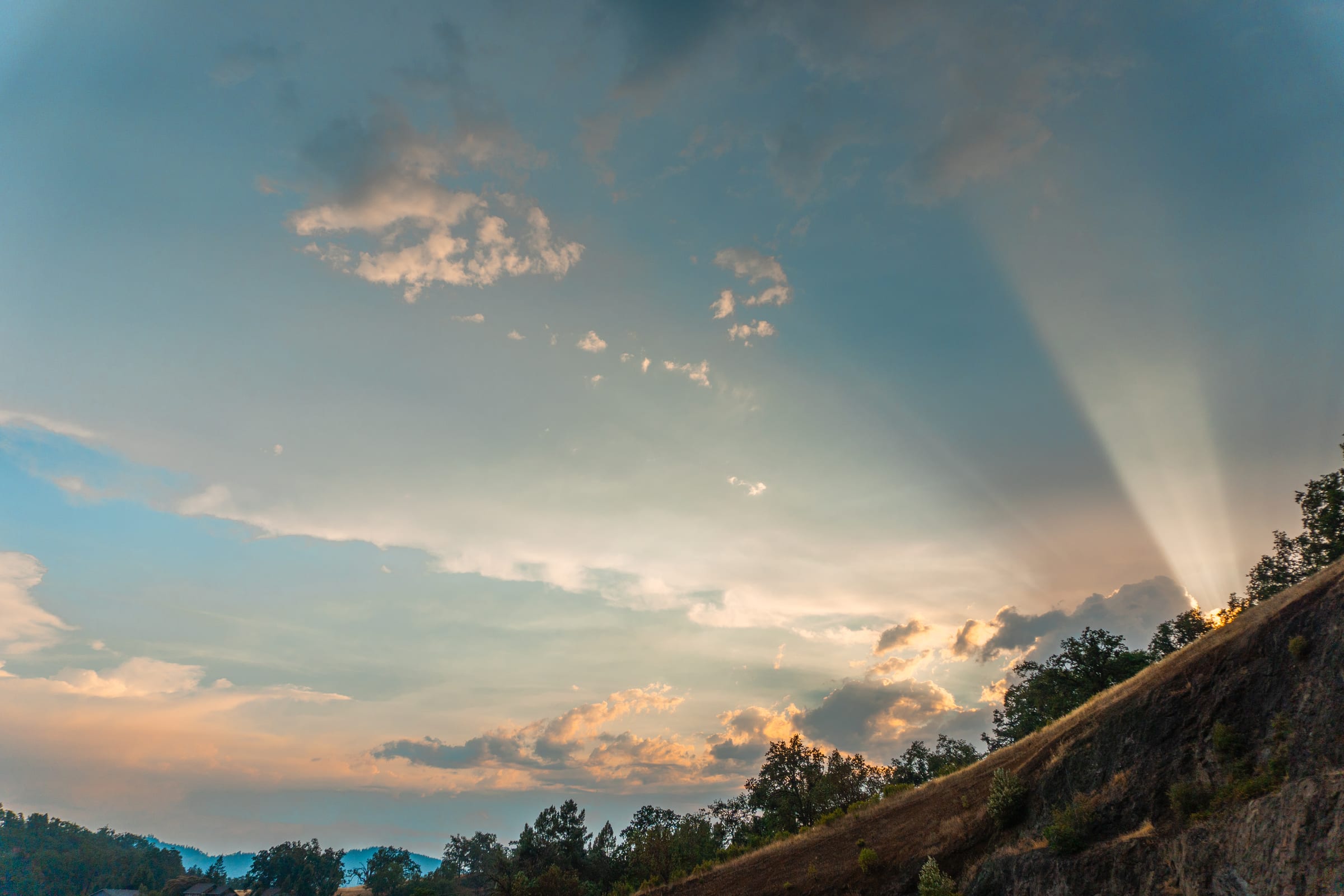 Rays of July sunset light fanning across the clouds above a dark hillside at Melrose