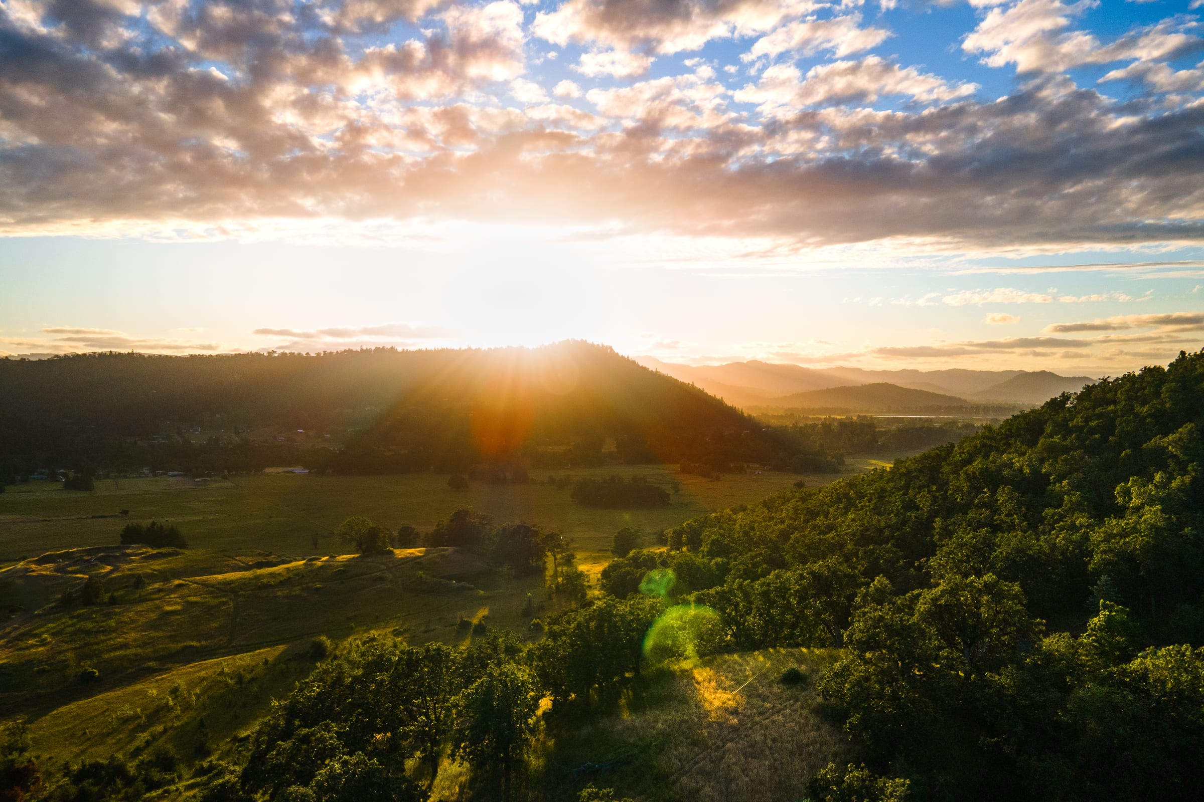 The June sun setting behind a hill at Roseburg, light spilling across fields and oaks