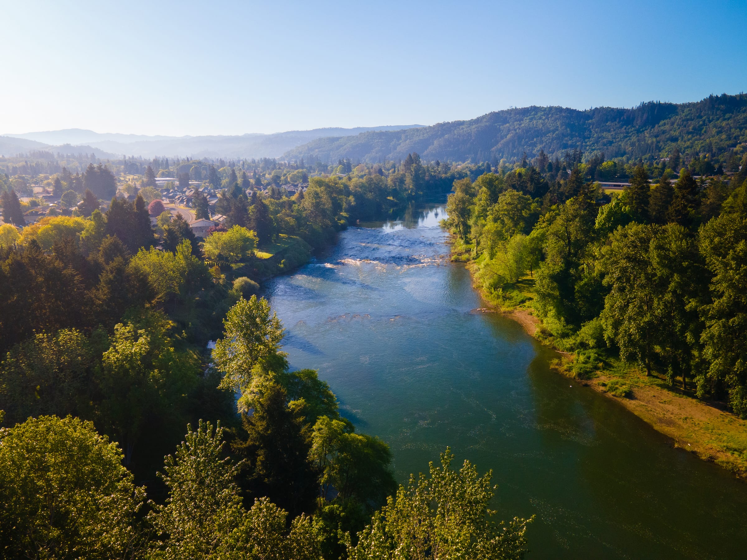 The South Umpqua River running between green trees and houses in Roseburg in May, wooded hills beyond