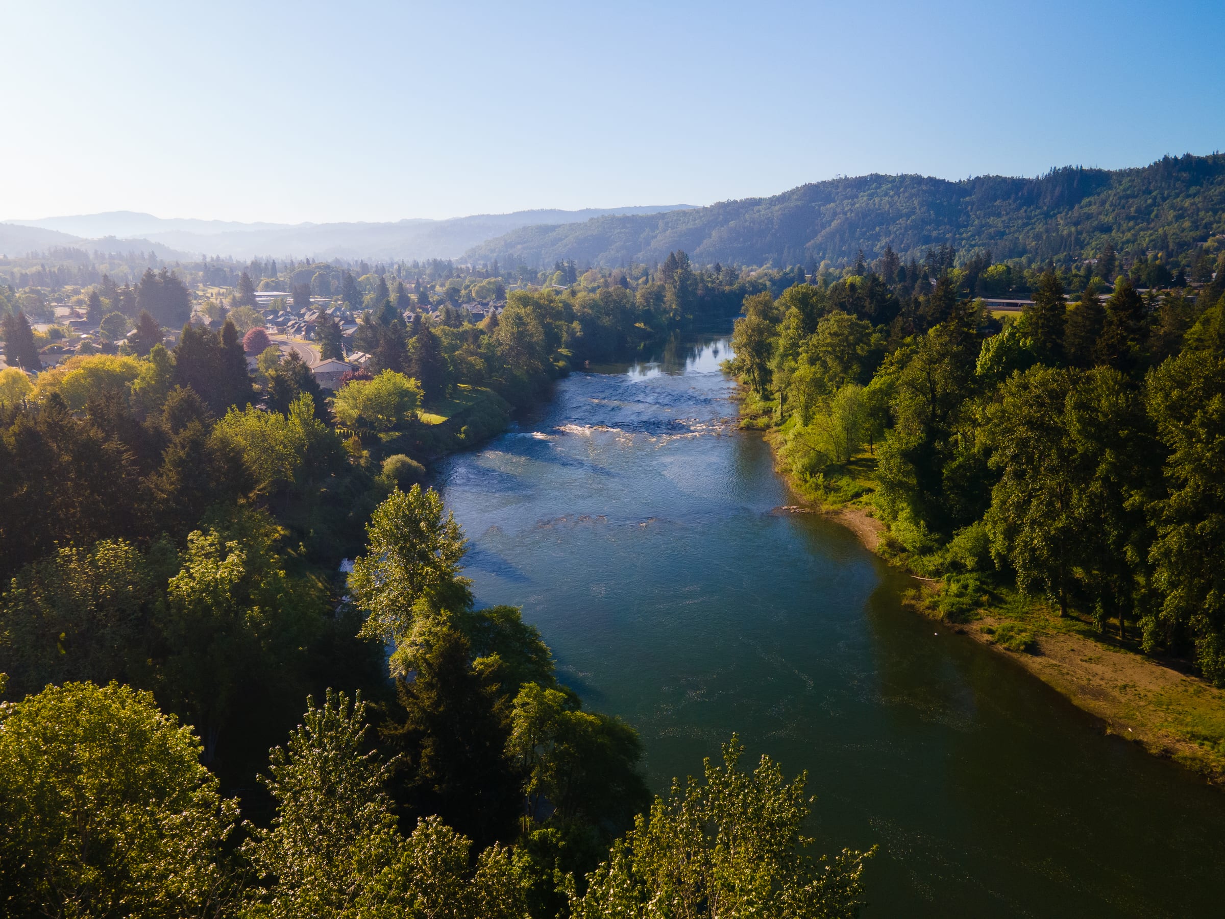 The South Umpqua River running past green trees and houses in Roseburg in May, wooded hills beyond